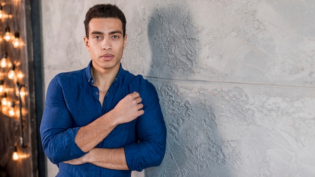 Retrato de un hombre joven hermoso elegante que mira la cámara que se inclina en la pared gris