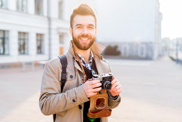 Retrato de un hombre joven hermoso sonriente que se coloca en la ciudad que sostiene la cámara disponible