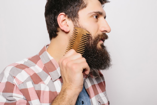 Retrato de hombre joven inconformista pelirroja atractiva con mirada seria y segura, sosteniendo el peine de madera y haciendo su espesa barba. Peluquero barbudo con estilo en camisa a cuadros peinando en el salón. Horizontal