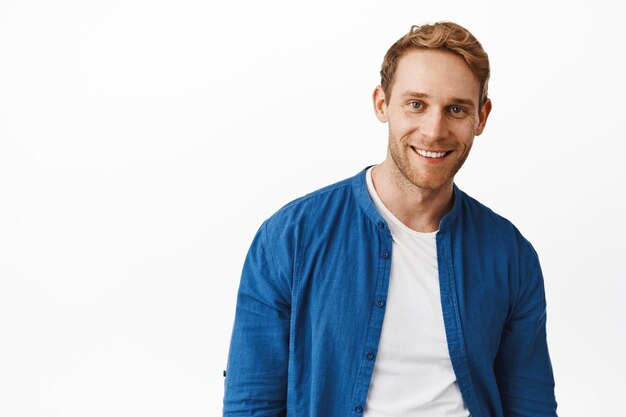 Retrato de hombre joven moderno guapo con cabello rojo y sonrisa blanca, mirando feliz y relajado a la cámara, de pie con camisa azul casual y camiseta contra el fondo del estudio