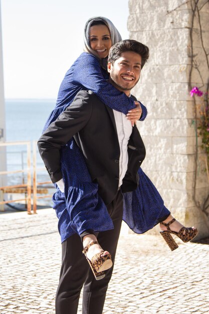 Retrato de hombre joven con mujer atractiva en la espalda. Sonriente hombre musulmán en traje negro con mujer en vestido de color detrás de él mientras la dama encantadora abrazando su cuello. Concepto de romance y relación