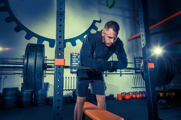 Retrato de hombre joven musculoso súper en forma trabajando en el gimnasio con barra en azul