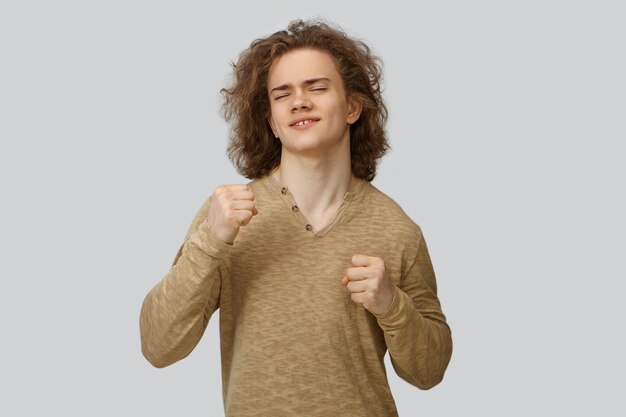 Retrato de hombre joven positivo guapo con cabello ondulado bailando, manteniendo los ojos cerrados. Atractivo chico sonriente emocional apretando los puños, expresando verdadera alegría, siendo feliz con buenas noticias