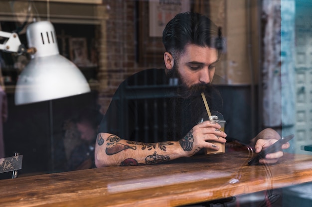 Retrato del hombre joven que bebe la leche del chocolate usando el teléfono móvil en café