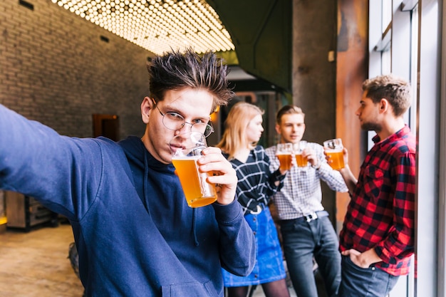 Retrato de un hombre joven que bebe el vaso de cerveza tomando selfie con sus amigos de pie en el fondo