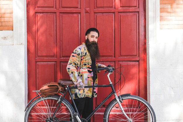 Retrato de un hombre joven que se coloca con la bicicleta delante de la puerta roja