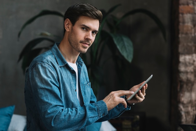 Retrato del hombre joven que mira que sostiene la tableta digital disponible que mira la cámara