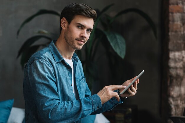 Retrato del hombre joven que mira que sostiene la tableta digital disponible que mira la cámara