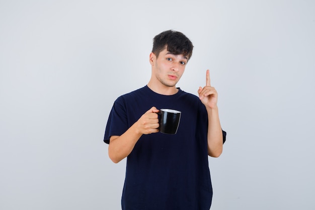 Retrato de hombre joven que muestra el gesto de eureka, apuntando hacia arriba, sosteniendo una taza de bebida en una camiseta negra y mirando la vista frontal inteligente