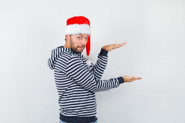 Retrato de hombre joven que muestra el signo de tamaño en la sudadera con capucha, gorro de Papá Noel y mirando vacilante