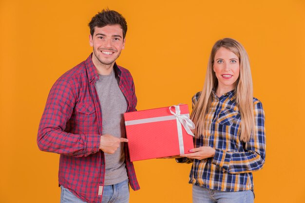 Retrato de un hombre joven que señala el dedo en el asimiento de la caja de regalo de su novia contra un fondo anaranjado