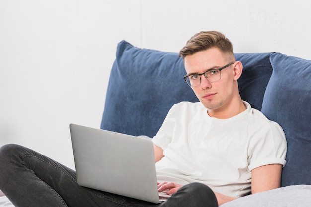 Retrato de un hombre joven que se sienta en cama con la computadora portátil que mira la cámara