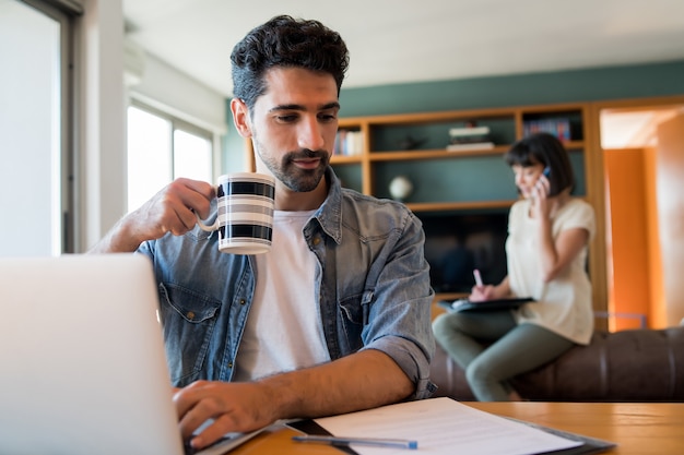 Retrato de hombre joven que trabaja con una computadora portátil desde casa mientras la mujer habla por teléfono