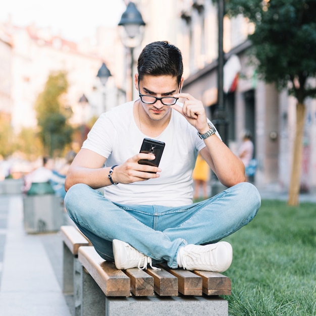 Foto gratuita retrato de un hombre joven sentado en el banco mirando la pantalla del teléfono inteligente