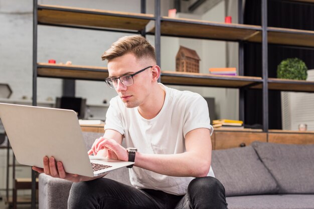 Retrato de un hombre joven serio que se sienta en el sofá usando la computadora portátil