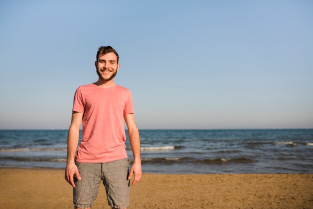 Retrato de un hombre joven sonriente que se coloca en la playa contra el cielo azul