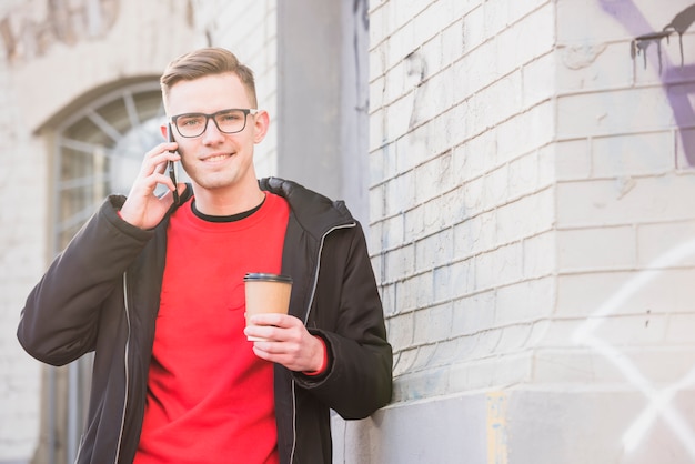 Retrato de un hombre joven sonriente que habla en el teléfono móvil que sostiene el café para llevar