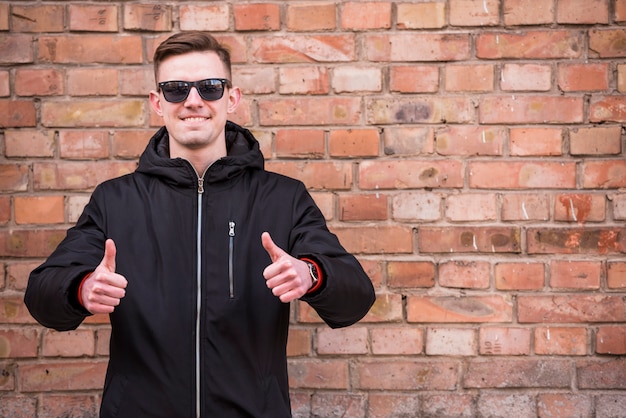 Retrato de un hombre joven sonriente que muestra el pulgar encima de la muestra que se opone a la pared de ladrillo