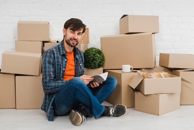 Retrato de un hombre joven sonriente que se sienta delante de las cajas de cartón que miran a la cámara