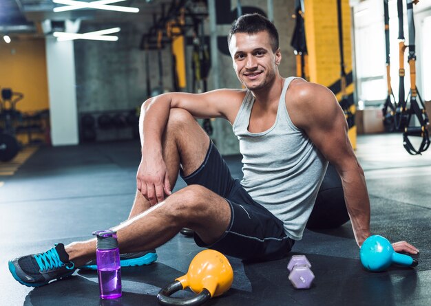 Retrato de un hombre joven sonriente que se sienta en piso cerca de equipos del ejercicio en gimnasio