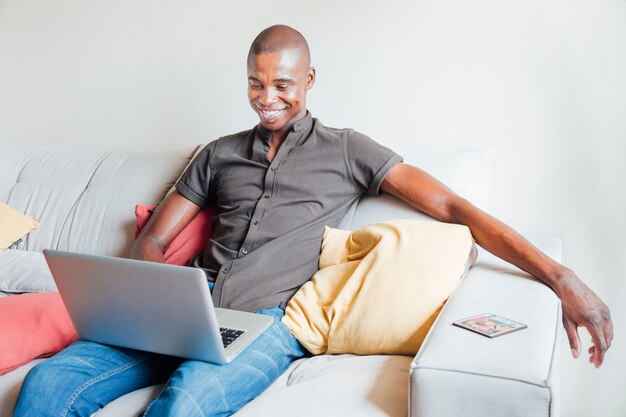 Retrato de un hombre joven sonriente que se sienta en el sofá usando la computadora portátil