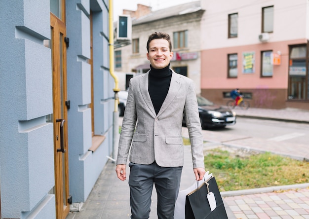 Retrato de un hombre joven sonriente que sostiene bolsos de compras que camina en la calle