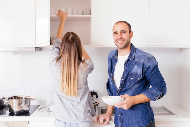 Foto gratuita retrato de un hombre joven sonriente que sostiene el cuenco en las manos que se colocan cerca de su esposa en la cocina