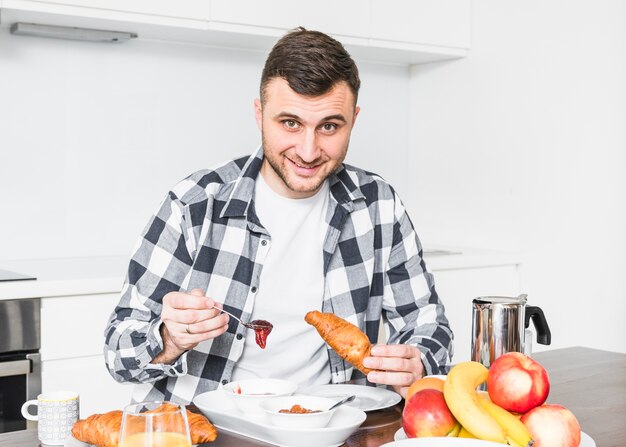 Retrato de hombre joven sonriente que tiene mermelada y croissant en mesa