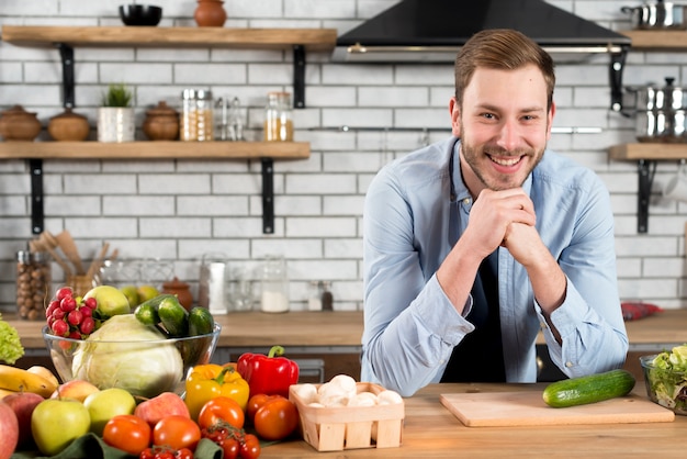 Retrato de un hombre joven sonriente con las verduras coloridas en la tabla en la cocina