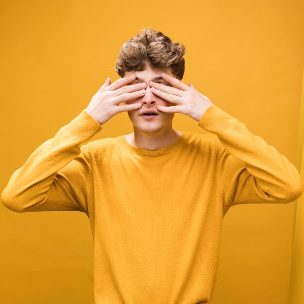 Retrato de hombre joven tapando sus ojos en un escenario amarillo