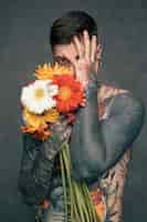 Foto gratuita el retrato de un hombre joven tatuado descamisado que sostiene el gerbera florece en la mano