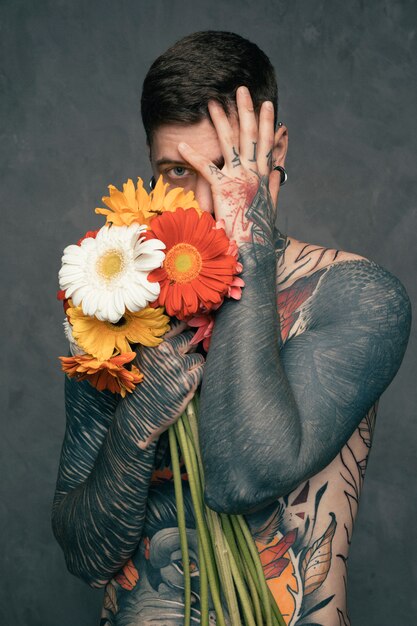 El retrato de un hombre joven tatuado descamisado que sostiene el gerbera florece en la mano