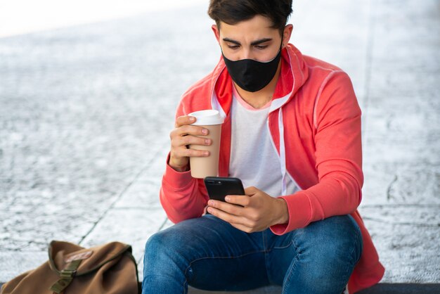 Retrato de hombre joven usando su teléfono móvil y tomando café mientras está sentado al aire libre en la calle. Hombre vestido con mascarilla. Concepto urbano.