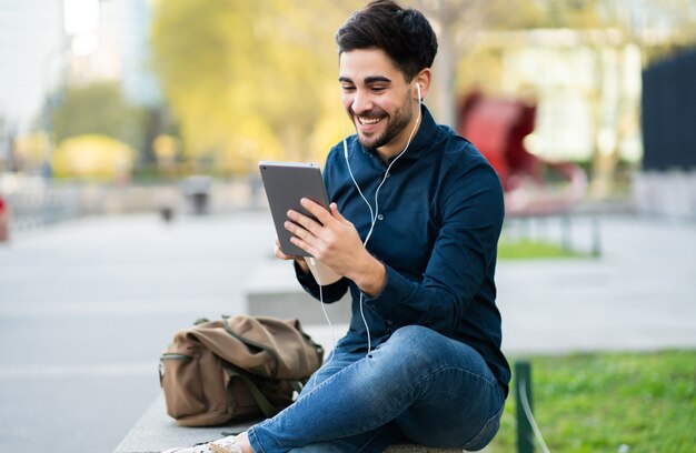 Retrato de hombre joven con una videollamada en tableta digital mientras está sentado en un banco al aire libre. Concepto urbano.