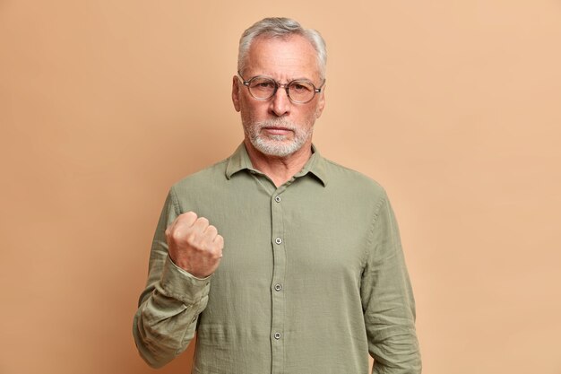 Retrato de hombre maduro barbudo irritado aprieta el puño y demuestra que la ira le advierte vestida con poses de camisa formal contra la pared beige