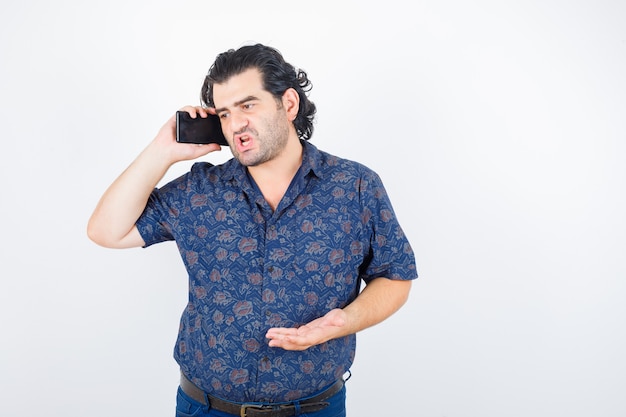 Retrato de hombre maduro, hablando por teléfono móvil en camisa y mirando enojado vista frontal