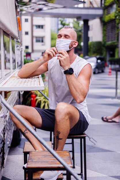 Retrato de hombre con máscara médica blanca en la plaza central de la ciudad se sienta en una silla en van cafe