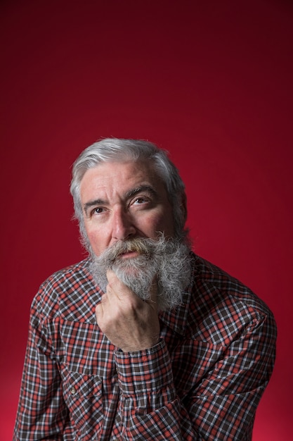 Foto gratuita retrato del hombre mayor contemplado con la mano en su barbilla que mira para arriba contra fondo rojo