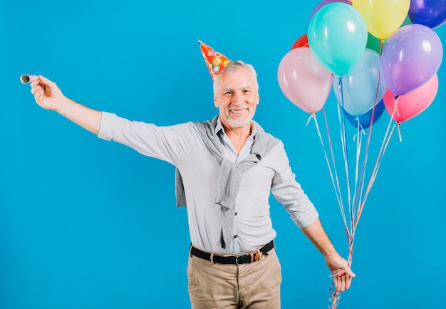 Retrato de un hombre mayor feliz que sostiene los globos y el cuerno del partido en el contexto azul