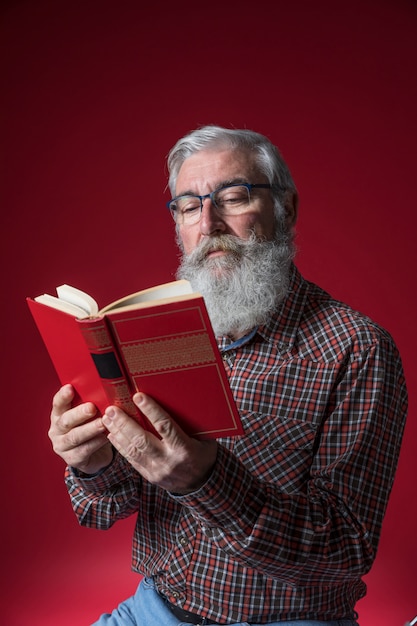 Retrato de un hombre mayor que lee el libro que se sostiene a disposición contra fondo rojo