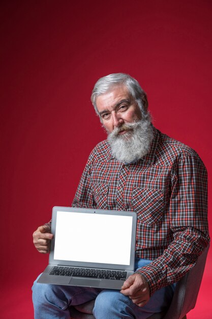 Retrato de un hombre mayor que se sienta en la silla que muestra el ordenador portátil con la pantalla blanca en blanco contra fondo rojo