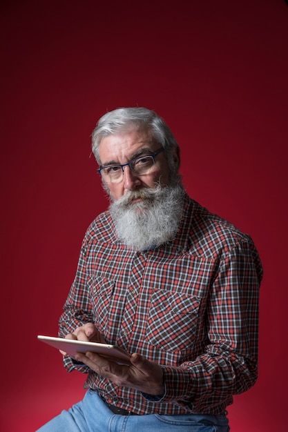 Foto gratuita retrato de un hombre mayor que sostiene la tableta digital en la mano que mira a la cámara contra el contexto rojo