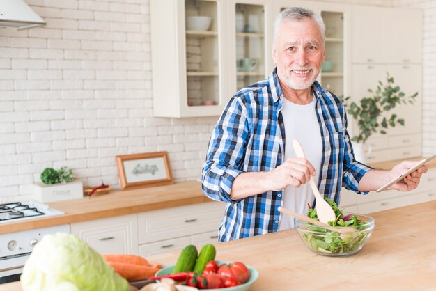 Retrato de un hombre mayor sonriente que sostiene la tableta digital en la mano que prepara la ensalada