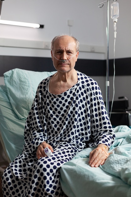 Retrato de un hombre mayor triste e indispuesto sentado en el borde de la cama del hospital con un goteo intravenoso adjunto y respirando con la ayuda de una máscara de oxígeno, mirando al frente
