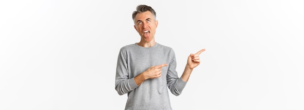 Foto gratuita retrato de un hombre de mediana edad decepcionado con el pelo gris que pone los ojos en blanco y muestra la lengua para expresar disgusto señalando con el dedo a la izquierda en una mala oferta promocional de pie sobre fondo blanco