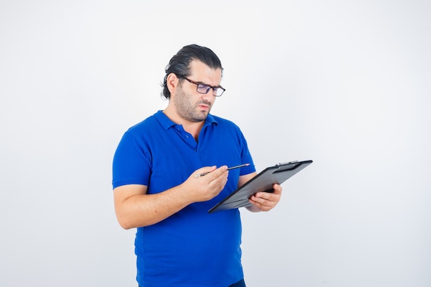 Retrato de hombre de mediana edad mirando el portapapeles mientras sostiene un lápiz en una camiseta de polo, gafas y mirando la vista frontal enfocada