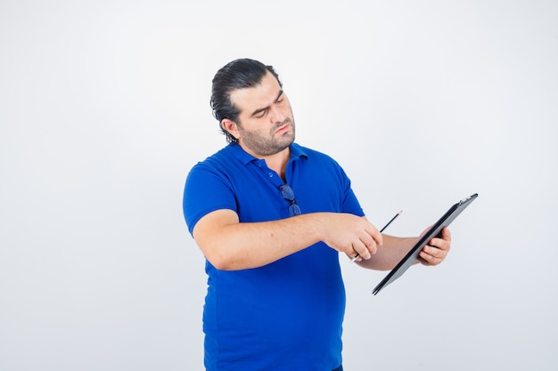 Foto gratuita retrato de hombre de mediana edad mirando a través del portapapeles mientras sostiene un lápiz en una camiseta de polo y mira pensativa vista frontal