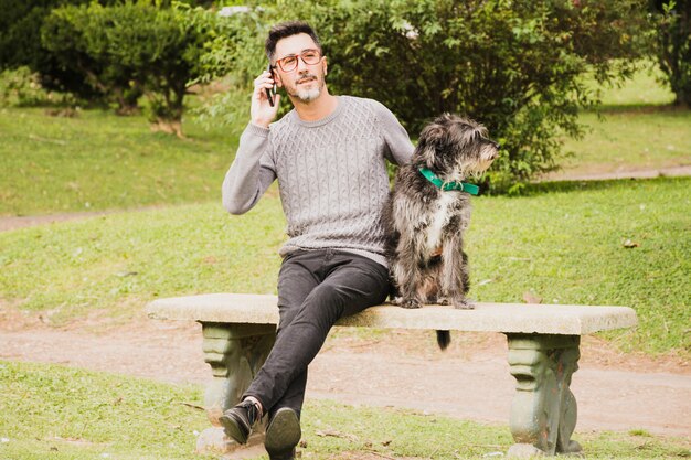 Retrato de un hombre moderno sentado en el parque con su perro hablando por teléfono móvil