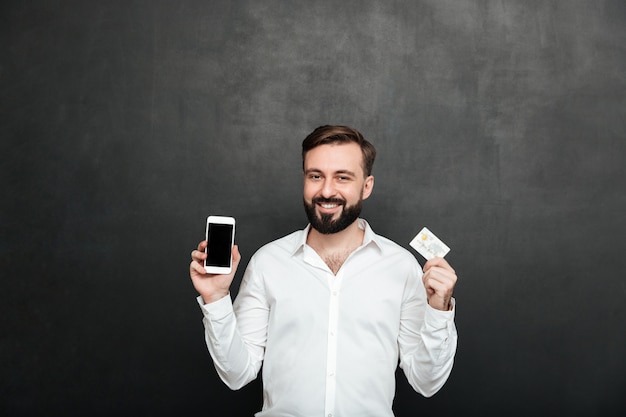 Retrato de hombre morena posando en la cámara con teléfono inteligente y tarjeta de crédito para compras en línea, aislado sobre gris oscuro