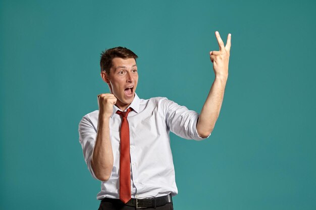 Retrato de un hombre moreno alegre con ojos marrones, vestido con una clásica camisa blanca y corbata roja. Está actuando como si hubiera ganado algo posando en un estudio sobre un fondo azul. Concepto de gesticular